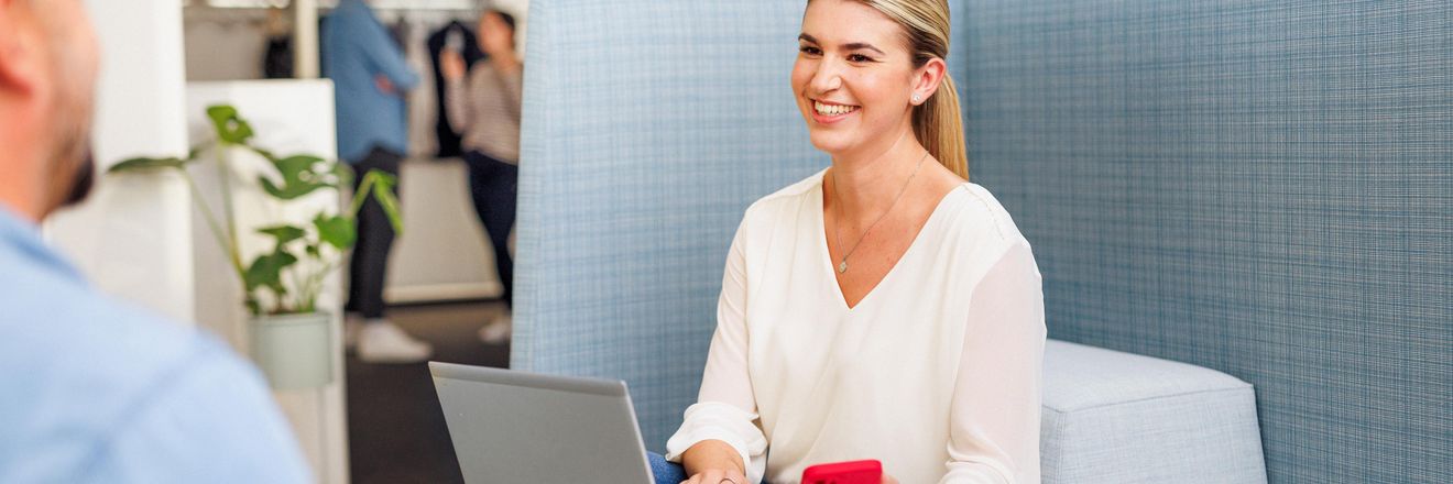 Kaufand-Mitarbeiterin in stylischer Büro-Lounge mit Laptop. Sie ist im Gespräch mit einem Kollegen.