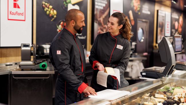 Eine Mitarbeiterin und ein Azubi in schwarzem Kaufland-Overall stehen hinter der Käsetheke. Die Mitarbeiterin erklärt etwas.