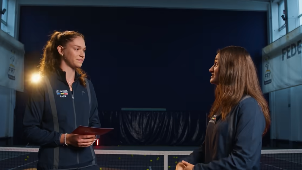 O imagine de interior, realizată pe un teren de tenis acoperit, ce surprinde două tinere într-o ipostază de interviu sau dialog. În partea stângă, o tânără cu părul șaten prins în coadă poartă o jachetă sport de culoare bleumarin, pe care este imprimat numele „GAE M.” și un mic steag al României; ea ține în mâini niște foi cartonate roșii și privește spre interlocutoarea ei. În partea dreaptă, a doua tânără, cu părul lung și lăsat liber, poartă o jachetă sport similară și vorbește activ, fiind surprinsă din profil.  În fundal se observă fileul de tenis, câteva mingi galbene împrăștiate pe teren și bannere publicitare alb-albastre ale Federației Române de Tenis (FRT). Lumina reflectoarelor din sală creează un efect de contur pe siluetele celor două fete.
