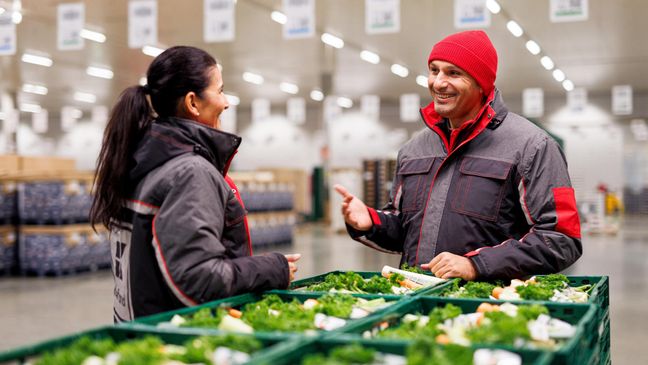 Ein Logistikmitarbeiter von Kaufland an gestapelten Gemüsekörben mit seiner Kollegin im Gespräch.