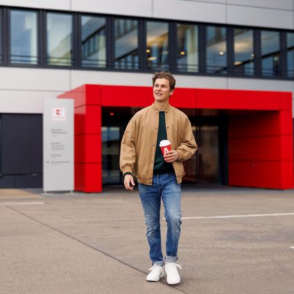 Ein Azubi, ein junger Mann in brauner Jacke, geht über den Platz vor dem Haupteingang der Kaufland-Zentrale.