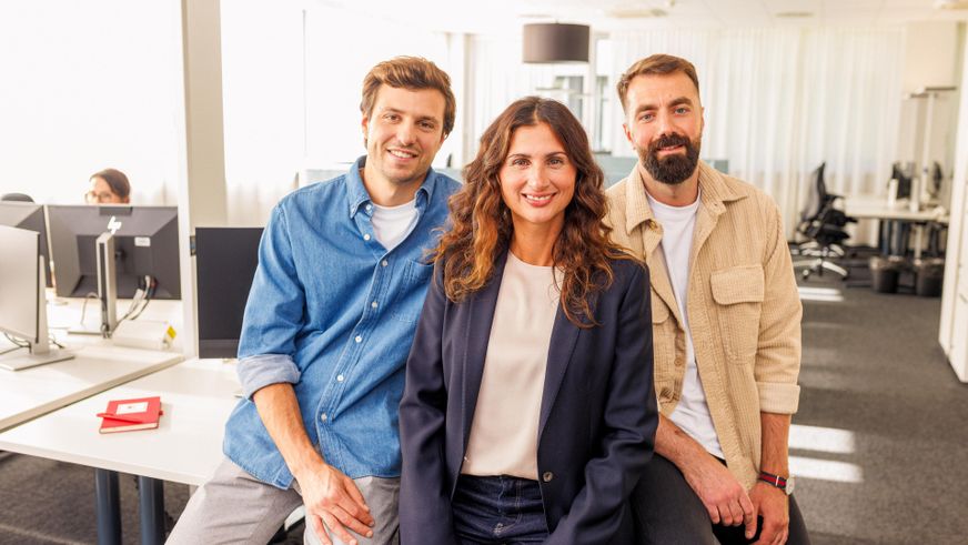 Großraumbüro: 3 Kaufland-Mitarbeiter posieren nebeneinander an der Kante eines Schreibtisches angelehnt für ein Gruppenfoto.