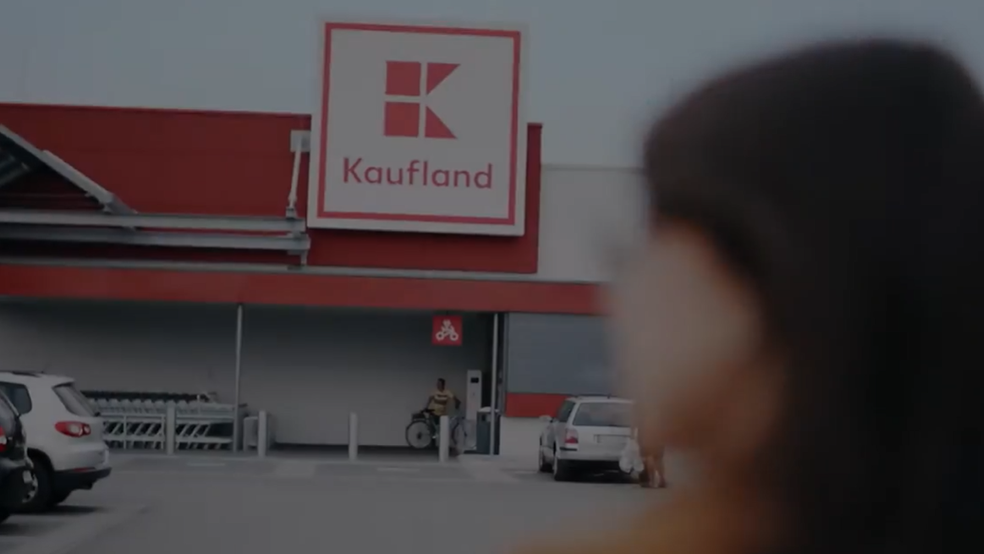 O imagine de la nivelul ochilor, ușor neclară, surprinde o parcare exterioară din fața unui magazin Kaufland, pe timp de zi, sub un cer înnorat.  În prim-plan, în partea dreaptă, se află ceafa și umărul drept ale unei persoane cu părul șaten închis, însă figura acesteia este complet blurată și scoasă din focar.  În fundal, clădirea magazinului domină centrul imaginii, având o fațadă roșie pe care este amplasat logo-ul mare, pătrat și alb al brandului Kaufland, cu litera „K” stilizată în roșu și numele scris dedesubt. Sub logo se află zona de intrare umbrită, unde se pot observa cărucioare de cumpărături aliniate și o persoană cu o bicicletă. În stânga și în centrul parcării de asfalt sunt staționate câteva autoturisme, printre care o mașină neagră și una albă în partea stângă, și o mașină argintie în dreptul intrării.