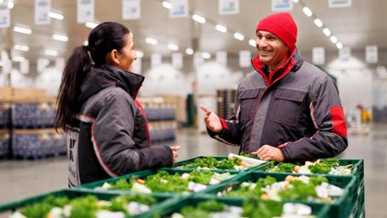 Ein Logistikmitarbeiter von Kaufland an gestapelten Gemüsekörben mit seiner Kollegin im Gespräch.