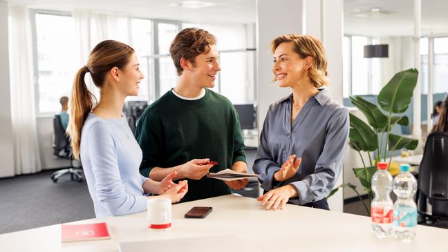 Kollaboratives Arbeiten mit einer weiblichen Führungskraft und zwei Azubis. Die drei stehen in legerer Kleidung am Hochtisch und unterhalten sich.