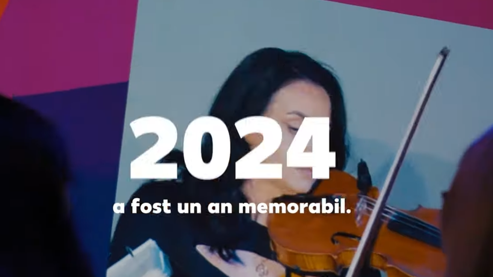 O imagine cadru dintr-o prezentare video, înfățișând o femeie cu părul brunet care cântă la vioară. Aceasta este concentrată, ținând arcușul pe corzi, iar fundalul din spatele ei este simplu, de culoare deschisă. În prim-plan, în părțile laterale stânga și dreapta, se observă siluetele neclare ale altor persoane din public. Peste imagine este suprapus un text alb, centrat: numărul „2024” scris foarte mare și îngroșat, iar dedesubt, cu litere mai mici, textul „a fost un an memorabil.”. În colțul din stânga sus apare un fragment dintr-un fundal grafic abstract în nuanțe de roz și mov.