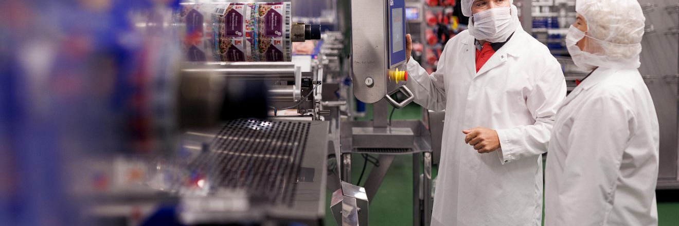 Fertigungsanlage in einem Kaufland-Fleischwerk. Zwei Kaufland-Mitarbeiter bedienen eine Produktionsanlage und unterhalten sich.