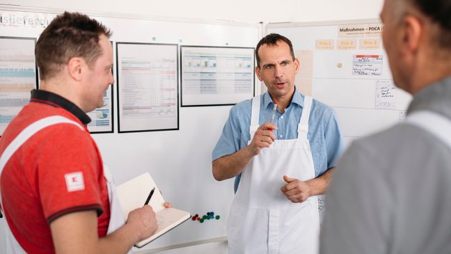 3 Kaufland-Fleischwerk-Mitarbeiter in der Besprechung. Ein Mitarbeiter macht sich Notizen.
