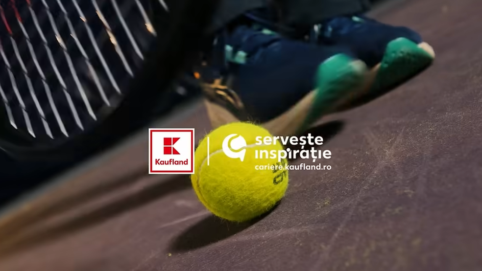 O imagine detaliată, realizată de la nivelul solului pe un teren de tenis, ce surprinde o minge de tenis galben-verzuie aflată pe suprafața de joc de culoare vișinie. În stânga mingii este poziționat logo-ul pătrat „Kaufland”, iar în dreapta ei apare un element grafic alb însoțit de textul „servește inspirație” și adresa web „cariere.kaufland.ro”.  În fundalul ușor estompat (out of focus) se observă picioarele unui jucător încălțat cu pantofi sport bleumarin cu talpă verde, prinși în mișcare, iar în colțul din stânga sus apare o porțiune din racordajul unei rachete de tenis.