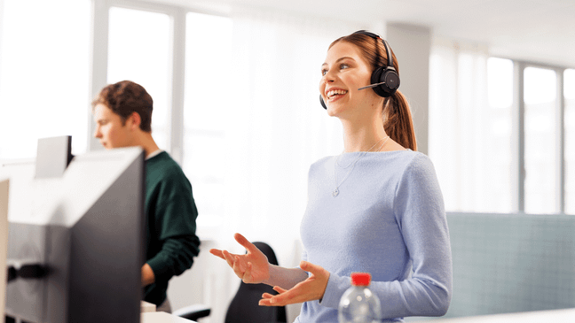 Ein weiblicher Azubi in hellblauem Pullover hat ein Headset auf und führt im Stehen ein Gespräch. Im Hintergrund sieht man halb verdeckt von einem Computer einen weiteren Azubi.
