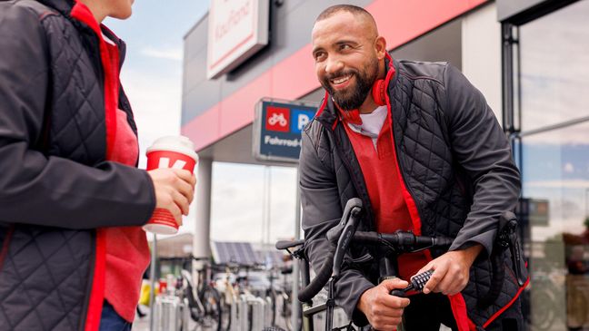 Pause vor der Kaufland-Filiale: Kaufland-Verkaufsmitarbeiter schließt sein Fahrrad an. Neben ihm eine Kollegin mit einem To-Go-Getränk bei der Pause.