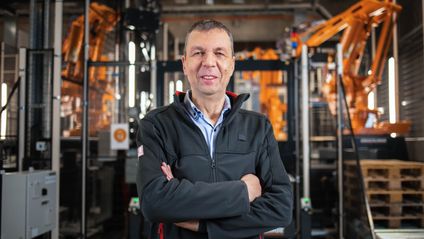 Teamleiter für Automatisierung am Logistikzentrum Geisenfeld steht mit verschränkten Armen vor einer Roboteranlage und lächelt freundlich in die Kamera