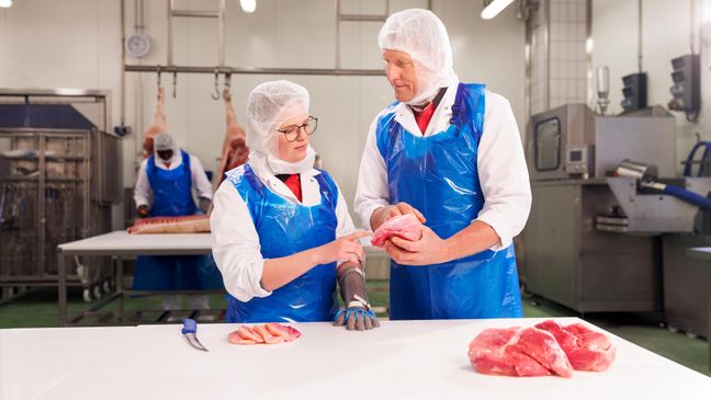 Ein Ausbilder und ein Azubi in blau-weißer Schutzkleidung stehen an einem Tisch in der Azubi-Werkstatt für die Fleischzubereitung. Die Azubi prüft Fleischqualität, während der Ausbilder ihr etwas erklärt.
