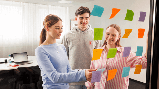 Zwei junge Frauen und ein junger Mann in legerer Kleidung stehen vor einer gläsernen Wand und arbeiten mit Post-Its an einem Projektboard.
