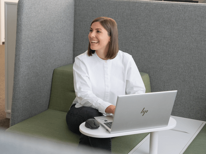 Projektleiterin Patricia aus dem Immobilienbereich sitzt in einer Sitzecke im Büro am Laptop und lacht zur Seite.