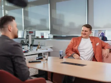 O imagine de interior, realizată într-un birou modern și luminos, ce surprinde o discuție sau un interviu între doi bărbați așezați la o masă de conferință din lemn deschis. În partea dreaptă, un bărbat zâmbitor, îmbrăcat cu un sacou cărămiziu (corai) peste un tricou alb, stă relaxat pe o canapea roz și privește spre interlocutorul său. Are un microfon de tip lavalieră prins de tricou. În partea stângă, celălalt bărbat este fotografiat din spate/profil, purtând un sacou gri închis. Pe masă se află un pahar cu apă, un telefon și o doză de băutură. Fundalul prezintă un spațiu de birouri de tip open-space, cu ferestre mari acoperite parțial de jaluzele gri.