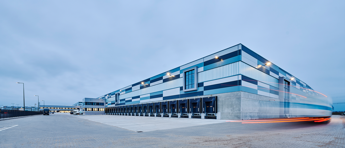 Zgrada logističkog centra u zoru, zablurano vozilo koje brzo prolazi pored zgrade.