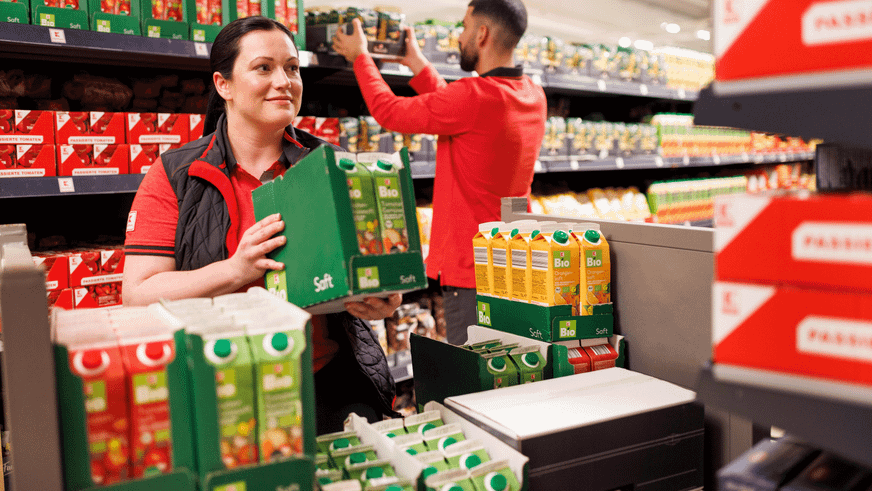 Ein Mitarbeiter und eine Mitarbeiterin stehen in einem Gang in einer Kaufland-Filiale. Die junge Frau ist von vorne zu sehen, der junge Mann von hinten. Beide räumen Ware ein.