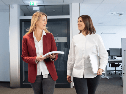 Zwei Frauen laufen einen Gang im Büro entlang und schauen sich nett an.