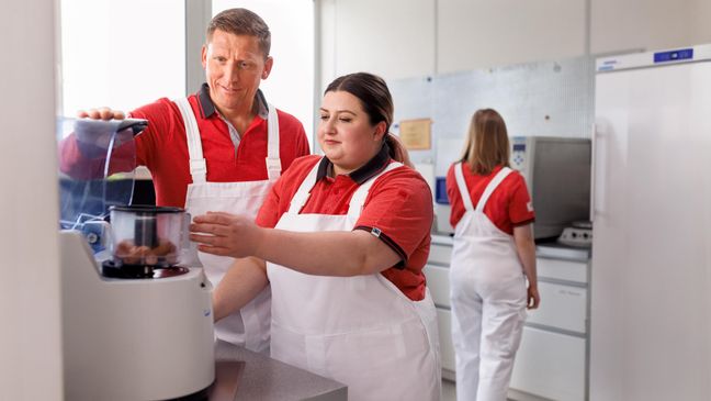 Im Qualitätsssicherungs-Labor steht eine Nachwuchskraft neben einem Ausbilder. Die beiden tragen eine weiße Latzhose und einen roten Pullover. Die junge Frau zeigt dem Ausbilder etwas. Im Hintergrund dreht ihnen eine weitere Frau in derselben Kleidung den Rücken zu.