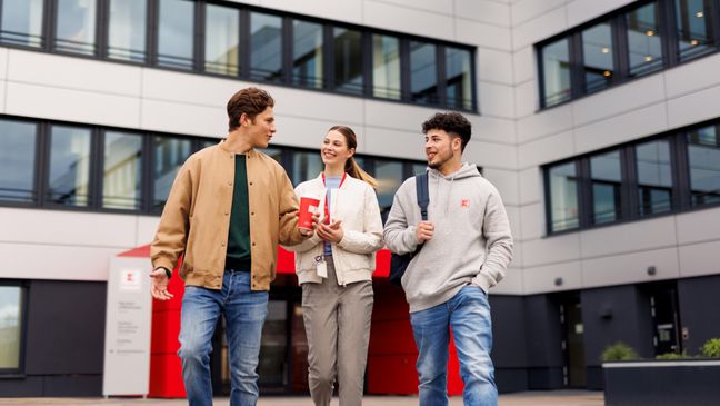 Drei Azubis stehen vor dem Haupteingang der Kaufland-Zentrale. Außen stehen zwei jungen Männer, in der Mitte eine junge Frau. Alle drei sind salopp gekleidet.