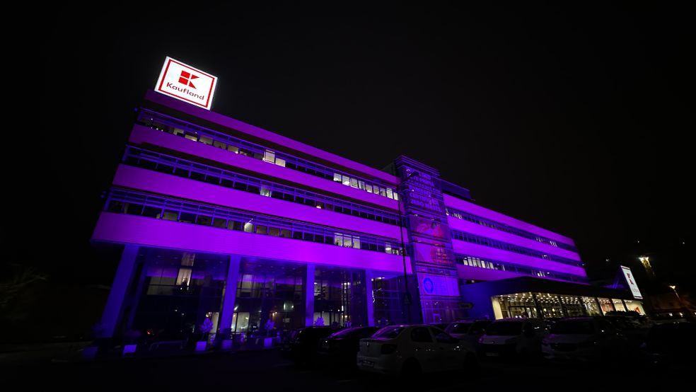 Clădirea sediului central Kaufland iluminată în culoarea mov pe timp de noapte, cu logo-ul luminos al companiei pe acoperiș și o parcare auto în față.