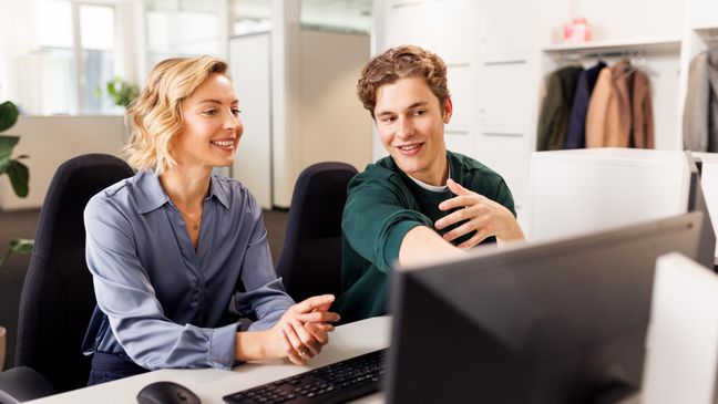 Eine Ausbilderin und ein Student sitzen am Arbeitsplatz vor einem Computer. Die beiden unterhalten sich.