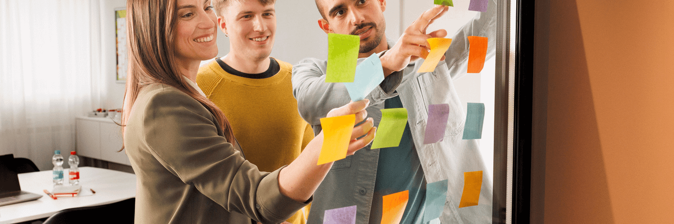 Eine junge Frau, ein junger Mann, beide Trainees und ein Ausbilder sind in einem Meeting. Die drei stehen vor einer gläsernen Wand und arbeiten mit Post-Its.